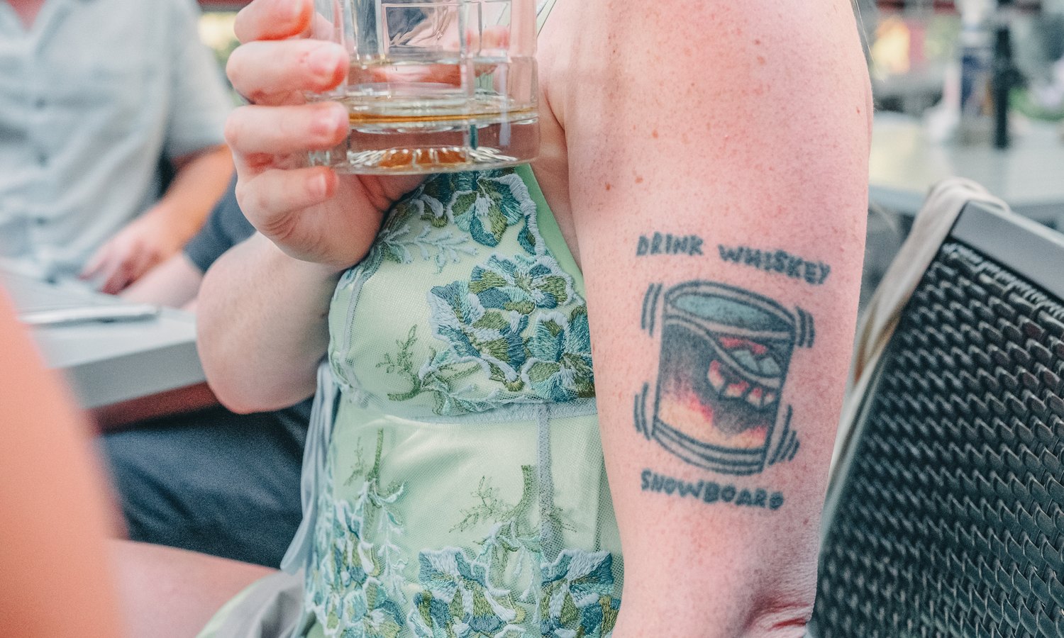 A guest on a sunlit patio holding a tumbler, a drink-whiskey-snowboard tattoo on her arm